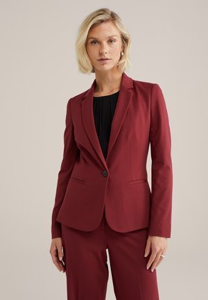 Červené na míru šitý blazer s single knoflíkovým zapínáním, klopovými límci a dvěma předními kapsami, doplněný o odpovídající kalhoty. Hladká textilie.