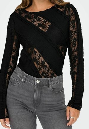 Vrouw draagt een zwarte bloemen kanten top met diagonale effen zwarte strepen en lange mouwen, gecombineerd met een grijze high-waisted jeans.