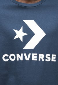 Μπλε σκούρο μπλουζάκι από βαμβακερό μείγμα υφάσματο ς με μεγάλο λευκό αστέρι και λογότυπο σήματος επάνω από τη λέξη "CONVERSE" με έντονα λευκά γράμματα.