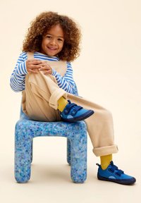 Scarpe blu con strappi in velcro, calze gialle, salopette beige e una camicia a maniche lunghe a righe, seduto su una sedia blu con macchie.