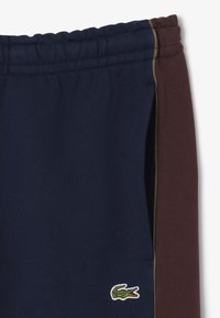 Pantaloni della tuta blu navy con cintura elastica, caratterizzati da un pannello laterale bordeaux e un logo ricamato verde vicino all'orlo. Tessuto morbido.
