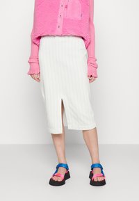 Jupe blanche, mi-longue, avec des rayures verticales et une fente à l'avant, associée à un pull rose texturé et des sandales multicolores à brides.