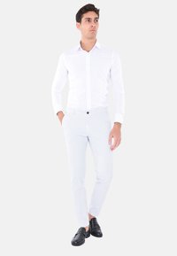 Camicia bianca a maniche lunghe, slim fit, con colletto a bottoni; abbinata a pantaloni slim fit grigi chiari; mocassini neri; sfondo neutro.