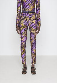 Lila outfit med en slim fit med gyllene barockmönster och svart text. Inkluderar långa ärmar och ett svart midjeband. Svarta sandaler.