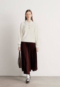 Biały sweter z dzianiny warkoczowej z kołnierzykiem i guzikami, zestawiony z długą, plisowaną, ciemnobrązową spódnicą. Do kompletu dodano brązową torebkę i czarne buty.