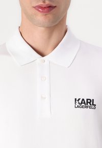 Fehér pólóing klasszikus gallérral, három gombbal és a bal oldalon fekete "KARL LAGERFELD" logóval.