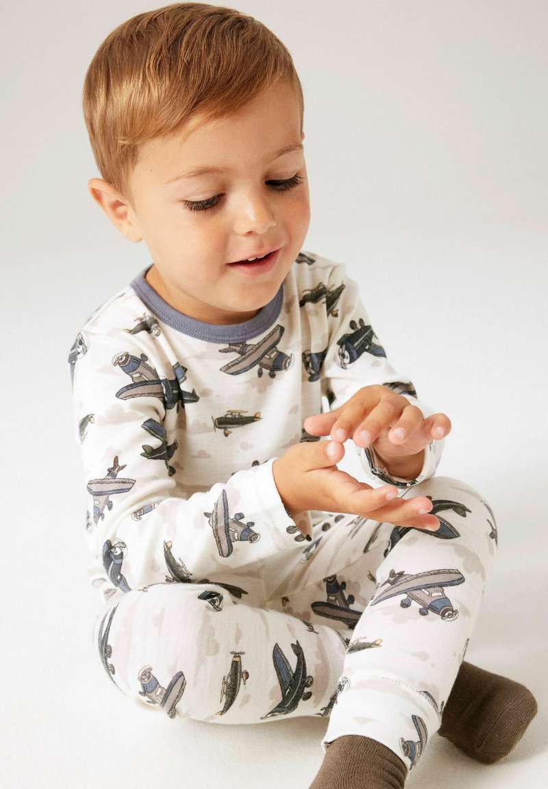 Ensemble de pyjama en coton avec motifs d'avions, couleurs claires, manches longues et poignets côtelés ; l'enfant est assis, les mains jointes.