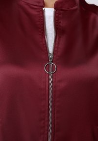 Μπουφάν από σατέν μπορντό με ψηλό γιακά, εμφανή ασημένιο φερμουάρ και κυκλικό τερματικό για το κλείσιμο, με λεία, γυαλιστερή υφή.