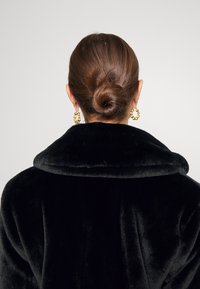 Femme aux cheveux bruns attachés en chignon, portant des boucles d'oreilles créoles dorées et un manteau en fourrure noire, vue de dos sur un fond uni.