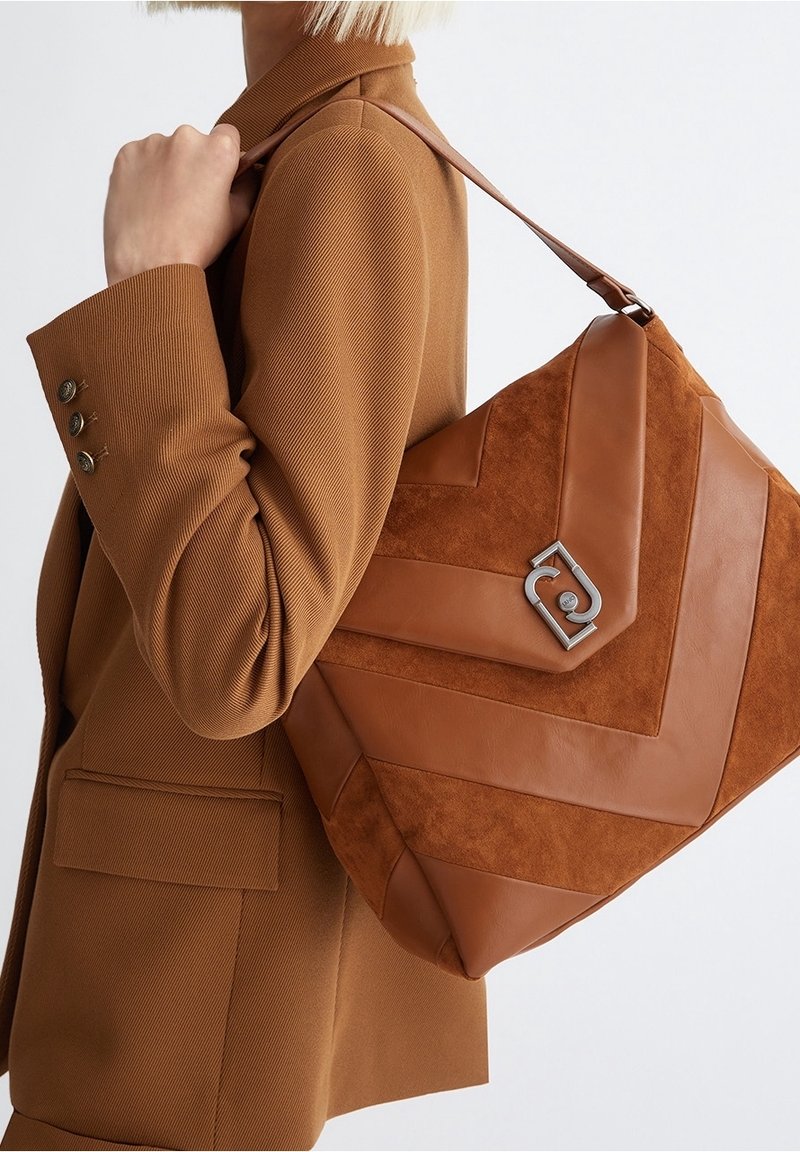 Femme en blazer marron portant un grand sac à main en daim et cuir couleur beige avec un motif géométrique et une fermeture argentée à l'épaule.