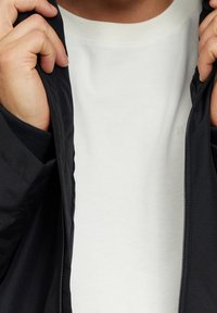Händer som drar upp en svart zip-jacka för att avslöja en enkel vit t-shirt under.