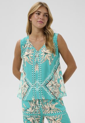 Femme portant un haut sans manches turquoise à motifs floraux et un pantalon assorti, debout les mains derrière le dos, regardant sur le côté.