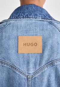 Gros plan sur le haut du dos d'une veste en jean bleue avec un patch en cuir beige portant l'étiquette HUGO cousu sous le col.