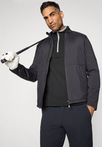Casaco insulated preto com fecho éclair, detalhes cinzentos, combinado com uma blusa preta de mangas longas e calças slim-fit; golfista segurando um taco.