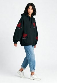 Sudadera negra con gráficos de corazones rojos y texto "SWEET". Presenta un bolsillo tipo canguro, un corte holgado, y se combina con jeans azul claro con puños y zapatillas blancas.