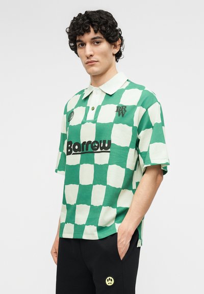 Νέος άντρας με σγουρά μαλλιά που φοράει πράσινο και λευκό καρό πόλο μπλουζάκι με την επιγραφή «Barrow» και μαύρο παντελόνι με ένα μικρό κίτρινο λογότυπο χαμογελαστής φατσούλας.