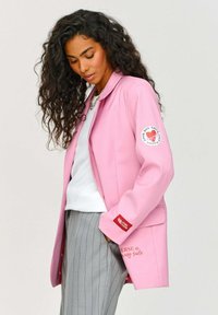 Růžová bunda s dlouhým rukávem, límcem a kapsou; má nášivku s logem na levém rukávu a potisk textu na boku. Nenosí se přes bílý top.