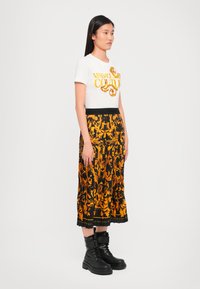 Bela majica s kratkimi rokavi z rumenim logotipom "Versace Jeans Couture", v kombinaciji s črno in oranžno vzorčeno nabrano krilo ter črnimi gležnjarji.