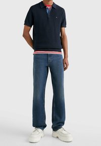 Marinblå texturerad poloskjorta med korta ärmar över en röd och vit randig skjorta, tillsammans med blå jeans och vita sneakers.