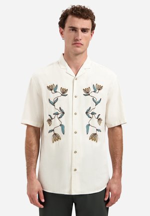 Mann trägt ein weißes kurzärmeliges Hemd mit bestickten blauen und gelben Blumenmustern auf der Vorderseite und steht vor einem einfarbigen Hintergrund.