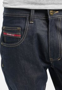 Jeans de mezclilla azul oscuro con costuras doradas, bolsillos delanteros y traseros, y un botón expuesto. Presenta una marca en rojo y blanco en el bolsillo trasero.