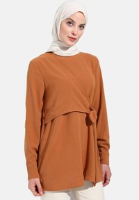 Haut à manches longues de couleur rouille avec un détail noué à la taille, fabriqué en tissu doux. Présente un col rond et une coupe décontractée.