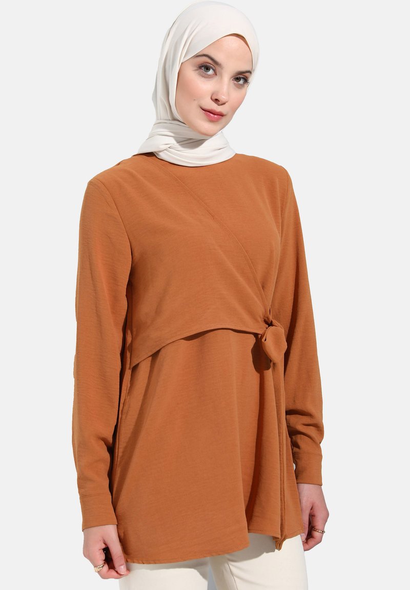 Haut à manches longues de couleur rouille avec un détail noué à la taille, fabriqué en tissu doux. Présente un col rond et une coupe décontractée.