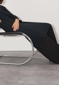 Černé látkové šaty sahající do poloviny lýtek v kombinaci s černými širokými kalhotami. Modelka se opírá o elegantní, zakřivenou židli z lesklého stříbrného kovu.