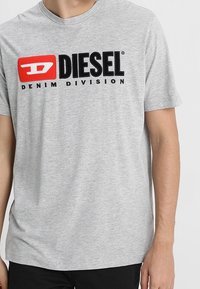 Šedé tričko s krátkým rukávem vyrobené z bavlny s kulatým výstřihem. Na přední straně má výrazné červené a černé logo "DIESEL DENIM DIVISION".