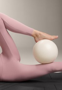 Rózsaszín leggings feküdnek egy fekete szőnyegen. Egy fehér, texturált edzőlabda pihen egy mezítláb alatt, bemutatva a felületén lévő hullámokat és mintákat.