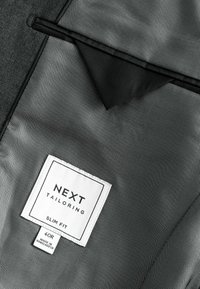 Szara siateczkowa tkanina z czarnym akcentem z nylonu posiada białą metkę z napisem "NEXT TAILORING", wskazującą na dopasowany krój, rozmiar 40R, wyprodukowaną w Bangladeszu.