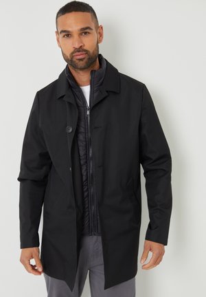 Abrigo negro tipo trench con un solo botón, superpuesto a una chaqueta acolchada negra con cremallera. Tejido suave, diseño ajustado y mangas largas.