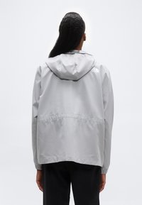 Άτομο που φα ίνεται από πίσω, φοράει ανοιχτό γκρι μπουφάν με κουκούλα και μαύρο παντελόνι, με σκηνικό απλό λευκό φόντο.