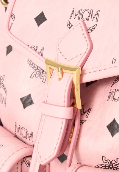 Gros plan sur la sangle en cuir rose d’un sac avec boucle dorée et motifs noirs du logo MCM sur un fond texturé.