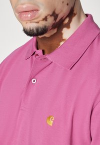 Gros plan sur une personne atteinte de vitiligo au niveau du cou et de la mâchoire, portant un polo Carhartt rose avec un logo jaune.
