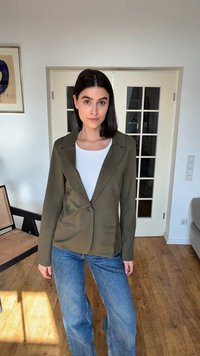 Γυναίκα με σκούρα μαλλιά που φοράει χακί σακάκι πάνω από λευκό τοπ και μπλε τζιν, στέκεται σε ένα φωτεινό δωμάτιο με ξύλινα πατώματα και πόρτες με γυάλινα πάνελ.