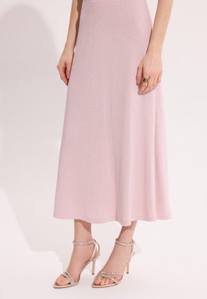 Kobieta w jasnoróżowej sukience midi o błyszczącej fakturze i beżowych sandałkach na wysokim obcasie z paskami ozdobionymi kryształkami, stojąca na jasnym tle.