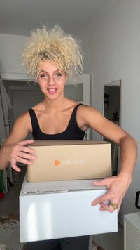 Una persona sostiene una caja marrón de Zalando sobre una caja blanca más grande. Ambas cajas son rectangulares y tienen logotipos impresos; la escena está bien iluminada.