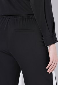 Svarta byxor med elastisk midja, med en slät yta och en bakfickdetalj. En knappfäst manschett synlig på armen.