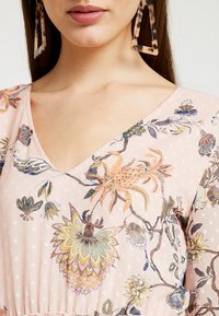 Roze jurk met bloemenpatroon, V-hals en getextureerde polka dots. Voorzien van ingewikkeld borduurwerk en geplooide details bij de taille.