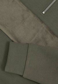 Tejido verde oliva con una sección suave y texturizada, presenta un zipper plateado. El diseño incluye variaciones de textil lisas y acanaladas.