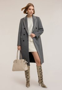 Cappotto di lana grigio con bottoni neri e rever a contrasto, abbinato a un top in maglia crema e una mini gonna bianca. Stivali con stampa serpente e borsa beige.