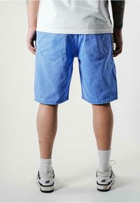 Shorts di jeans azzurri chiari con una vestibilità rilassata, dotati di due tasche posteriori, taglio dritto e una finitura leggermente sbiadita. Indossati con calzini bianchi e sneakers.