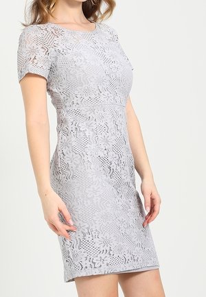 Frau trägt ein enganliegendes, kurzärmeliges, knielanges hellgraues Spitzenkleid mit floralen Mustern vor einfarbigem Hintergrund.