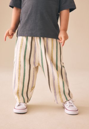 Barn iført sort skjorte, løse hvide bukser med lodrette grønne, gule, lilla og sorte striber og hvide sneakers, der står på beige gulv.