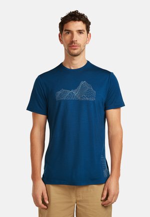 Man draagt een blauw T-shirt met korte mouwen en een geometrisch bergontwerp, gecombineerd met beige korte broek, staand tegen een effen witte achtergrond.