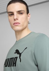 Vihreä puuvillapaita, jossa on suuri musta Puma-logo rinnassa. Lyhyet hihat ja pyöreä kaulus. Silkkinen rakenne.