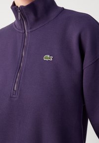 Paarse, getextureerde half-zip pullover gemaakt van zacht materiaal, met een klein groen Lacoste-logo op de borst.