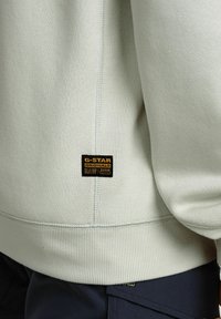 Felpa grigia chiara con texture a coste, dotata di un patch con il logo G-Star nero. Il design presenta cuciture piatte e una vestibilità rilassata.