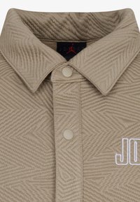 Šviesiai rudos spalvos tekstūruota marškinėliai su zigzago raštu, su apykakle, dviem sagomis ir baltu siuvinėtu "JORDAN" logotipu.